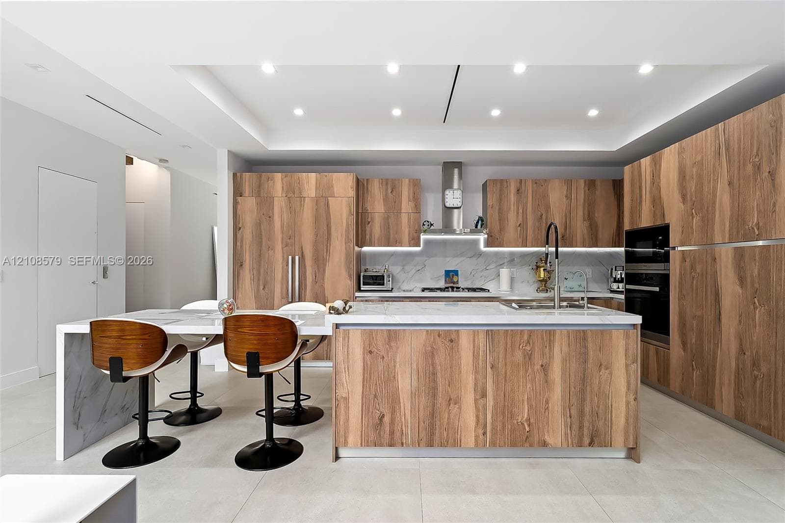 Kitchen featuring flat-panel wood-finish cabinetry, stone-surface countertops with waterfall edge, recessed ceiling lighting, built-in stainless steel appliances, and under-cabinet illumination