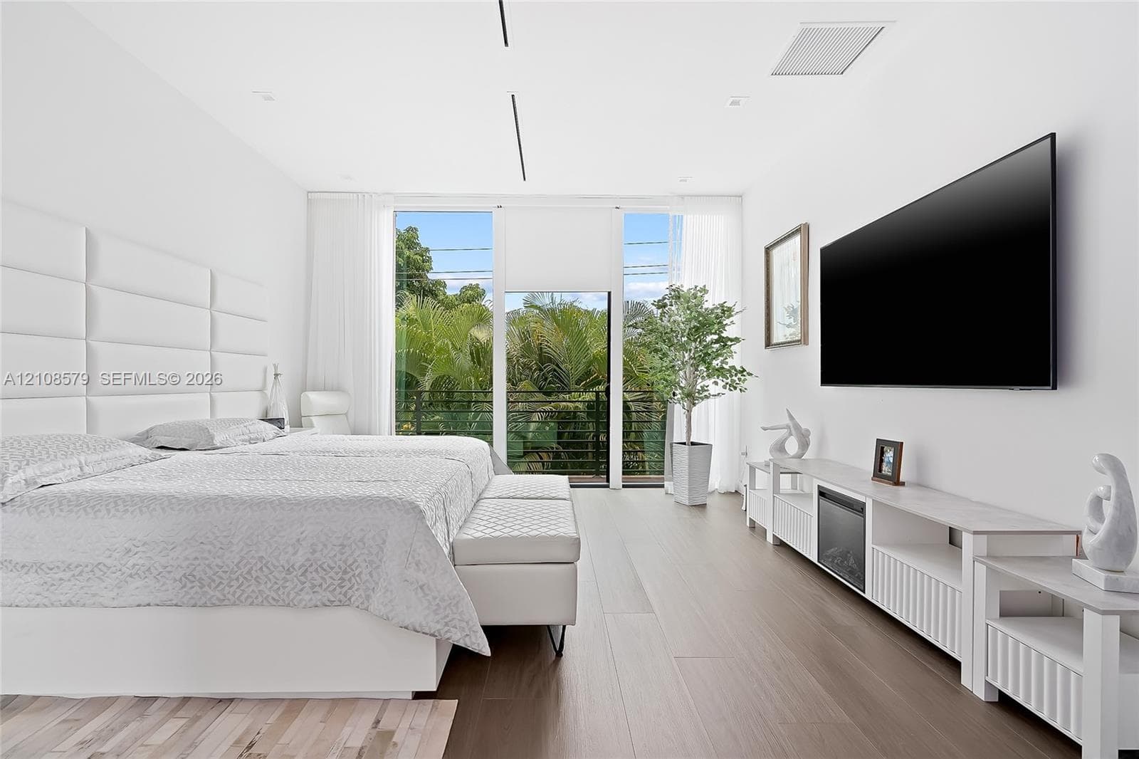 Wood-finish flooring, floor-to-ceiling windows with outdoor balcony access, and a built-in upholstered headboard wall paneling
