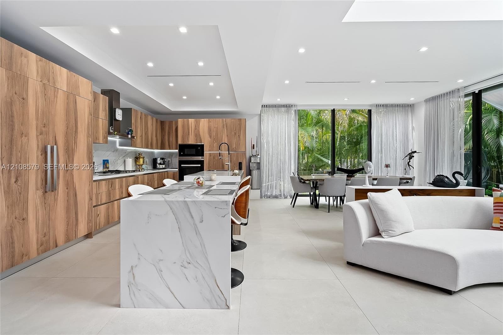 Kitchen featuring wood-finish cabinetry, stone-surfaced waterfall island, large-format tile flooring, recessed ceiling lighting, and floor-to-ceiling glass paneling