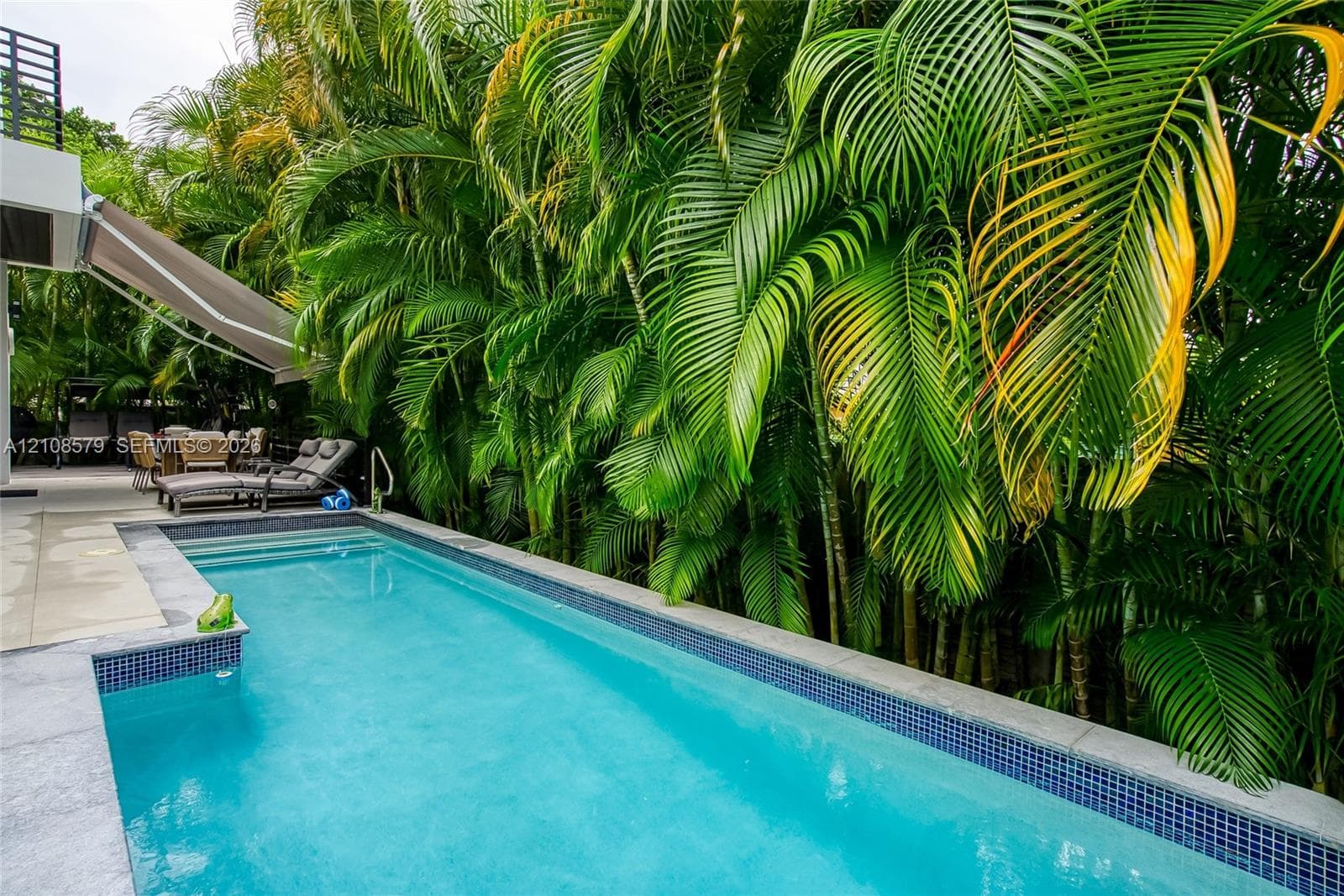 Rectangular swimming pool featuring tile waterline accents, integrated steps, a stone-finish pool deck, a retractable patio awning, and mature palm tree landscaping
