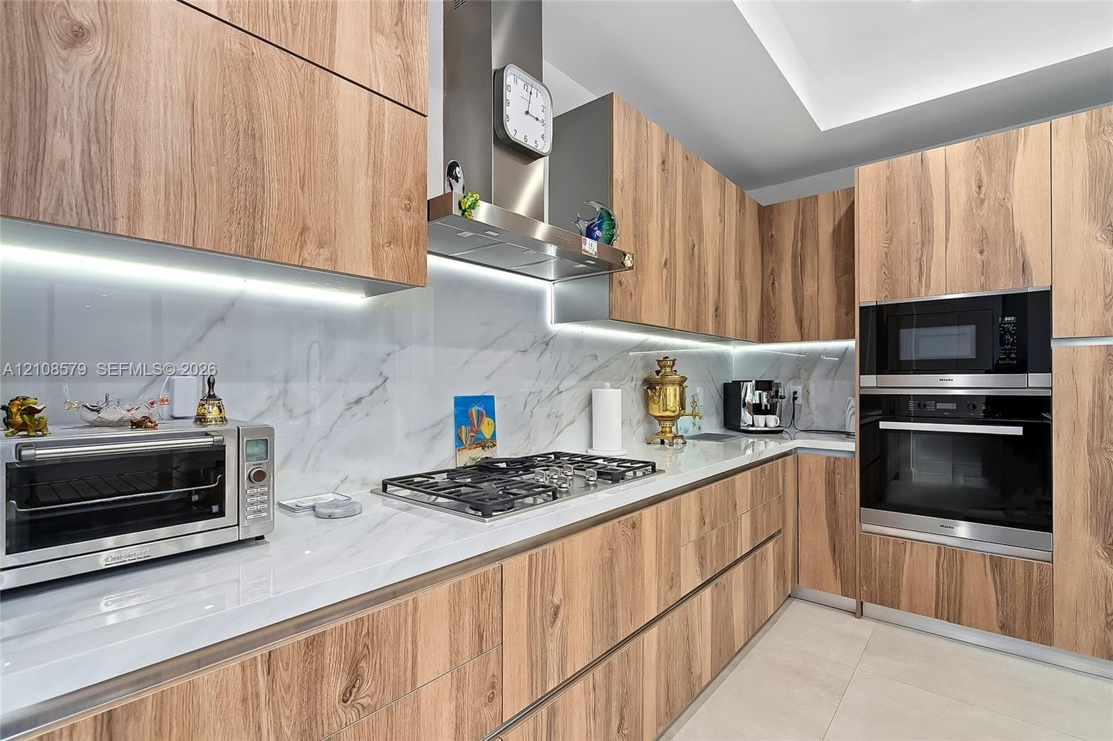 Kitchen featuring wood-finish cabinetry, stone-surface countertops, integrated under-cabinet lighting, a stainless steel gas cooktop with chimney-style range hood, and a built-in stainless steel oven and microwave combination