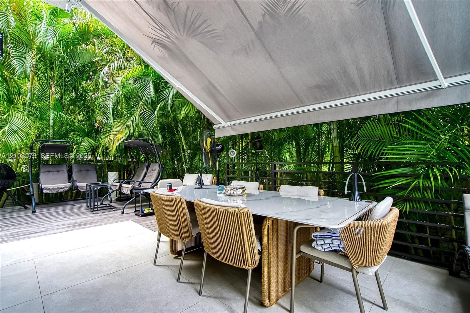 Outdoor terrace featuring large-format tile flooring, retractable fabric awning, and perimeter privacy fencing surrounded by mature tropical foliage