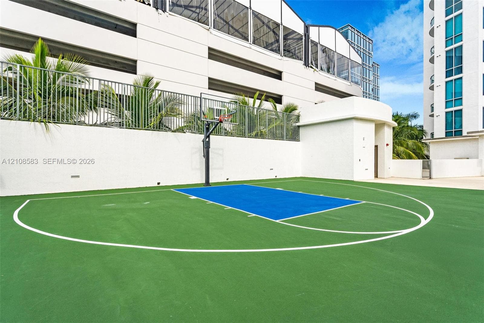 Outdoor basketball court with a multi-colored court surface, metal perimeter fencing, and integrated backboard system