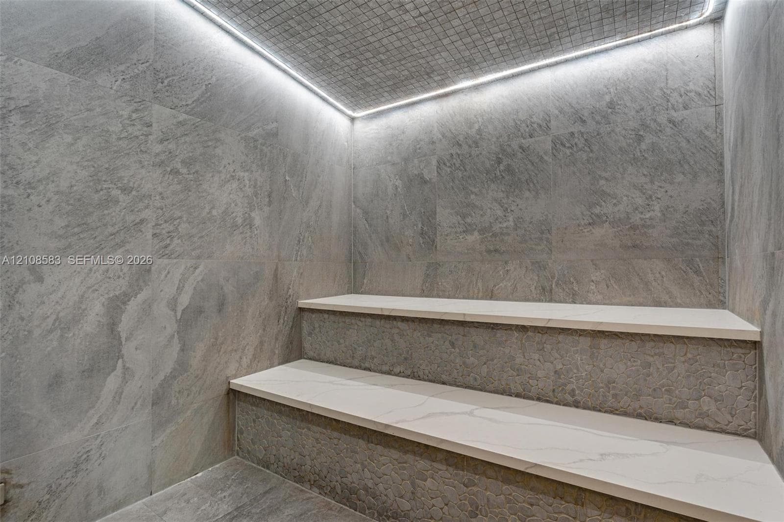Steam room featuring large-format stone-finish wall and floor tiling, multi-level tiered seating with smooth light-toned surfaces, textured stone-detail risers, and integrated perimeter LED cove lighting