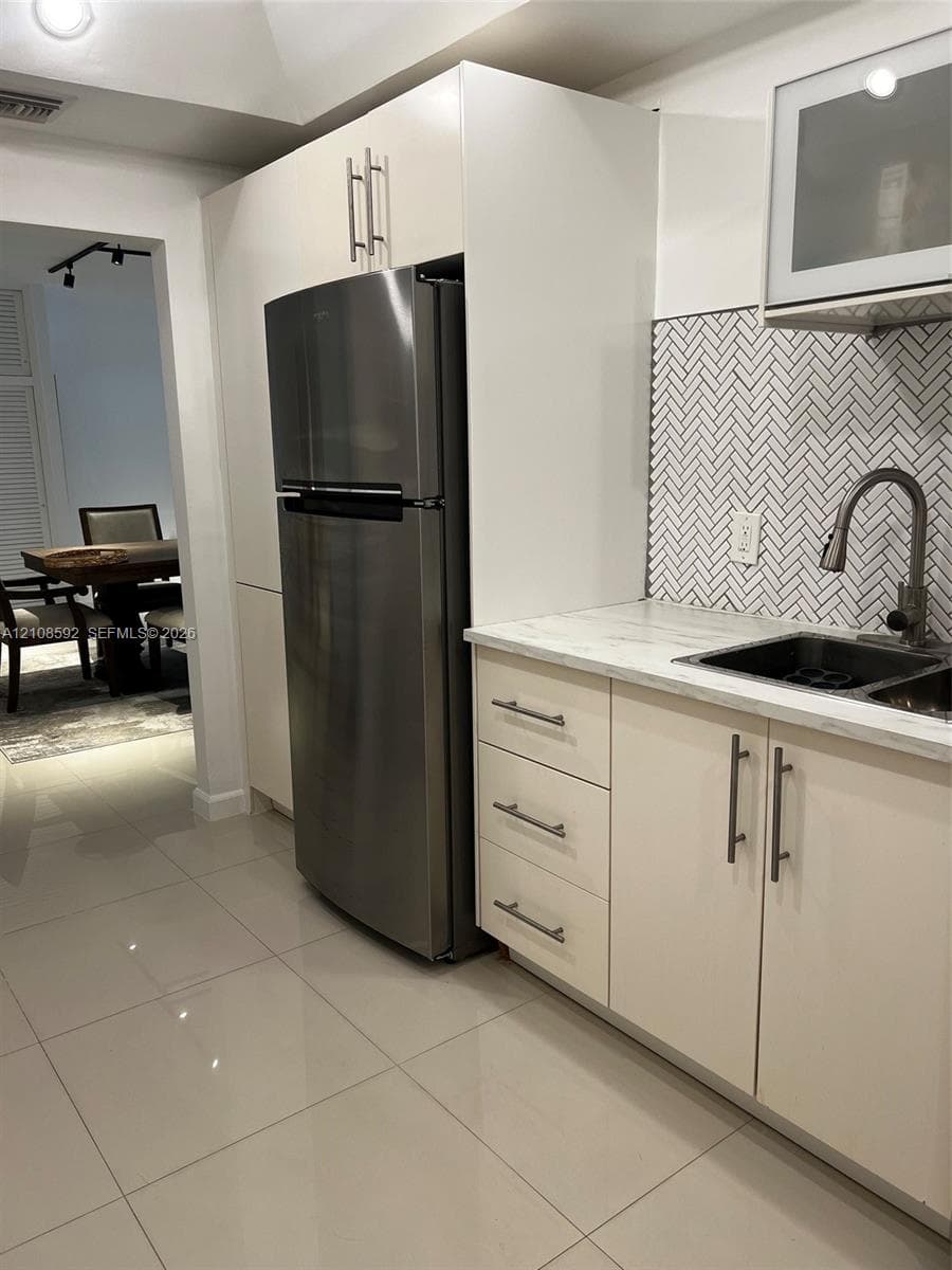 Kitchen featuring flat-panel cabinetry, stainless steel refrigerator, stone-surface countertop, herringbone tile backsplash, and undermount dual-basin sink with high-arc faucet
