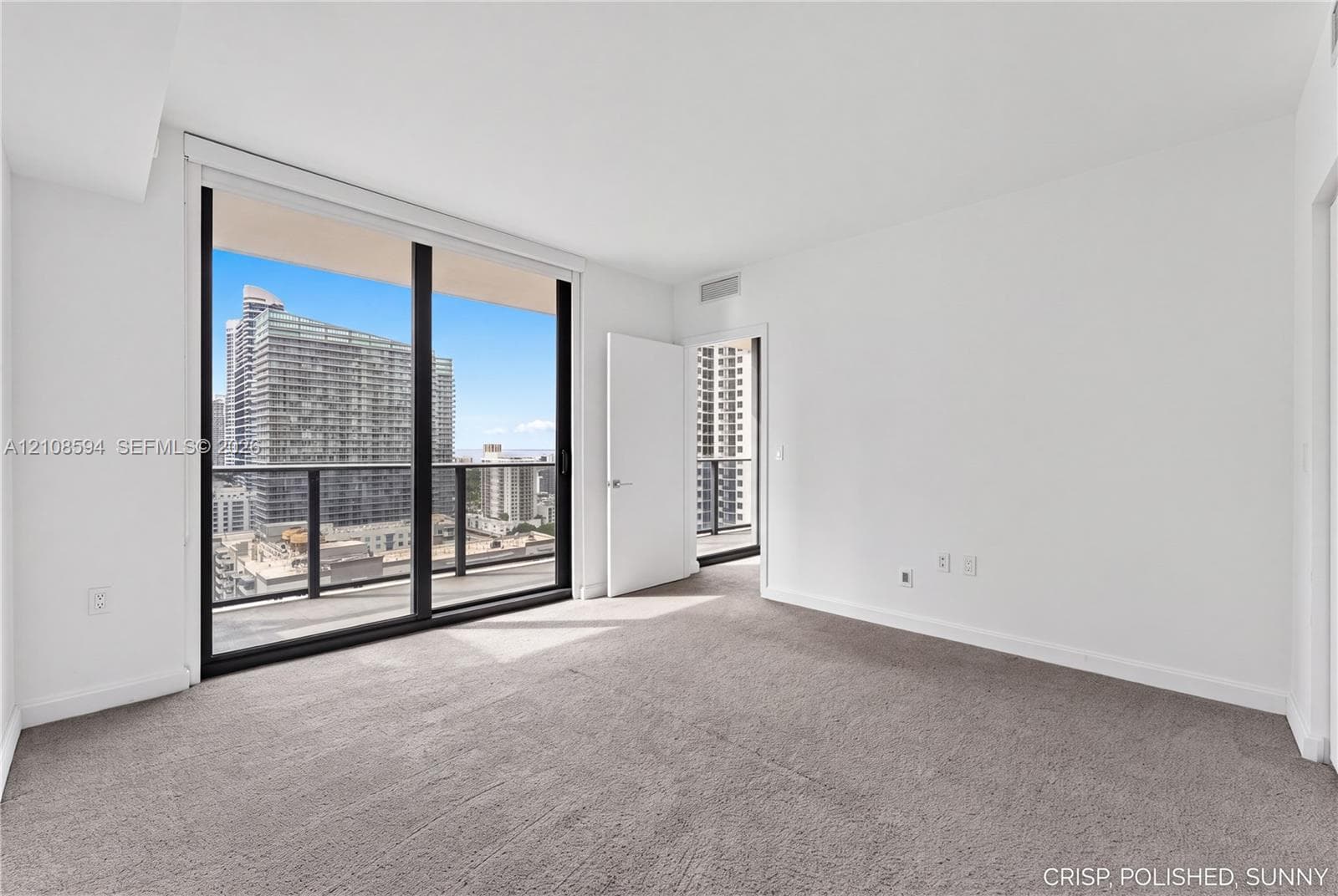 Floor-to-ceiling glass sliding doors, private balcony with metal railing, carpeted flooring, and urban cityscape scenery