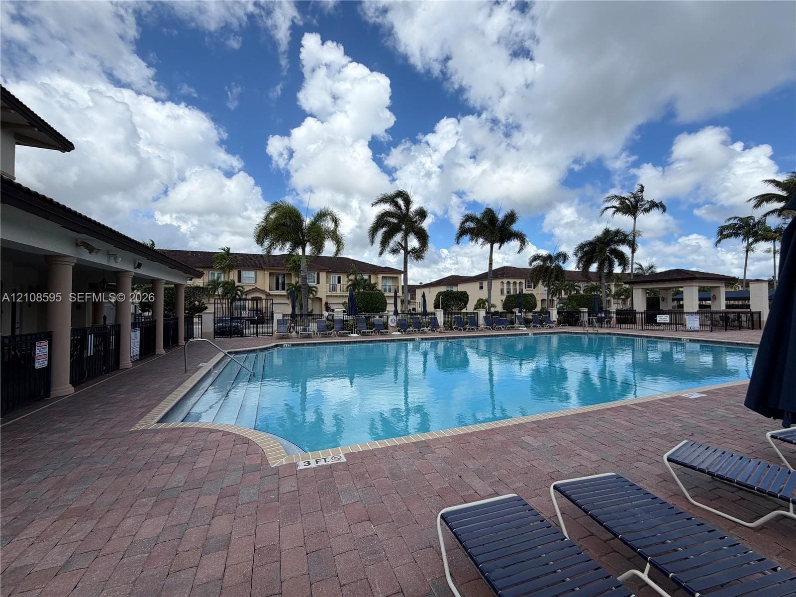 Community swimming pool featuring a brick paver deck, covered veranda with structural columns, and mature palm trees