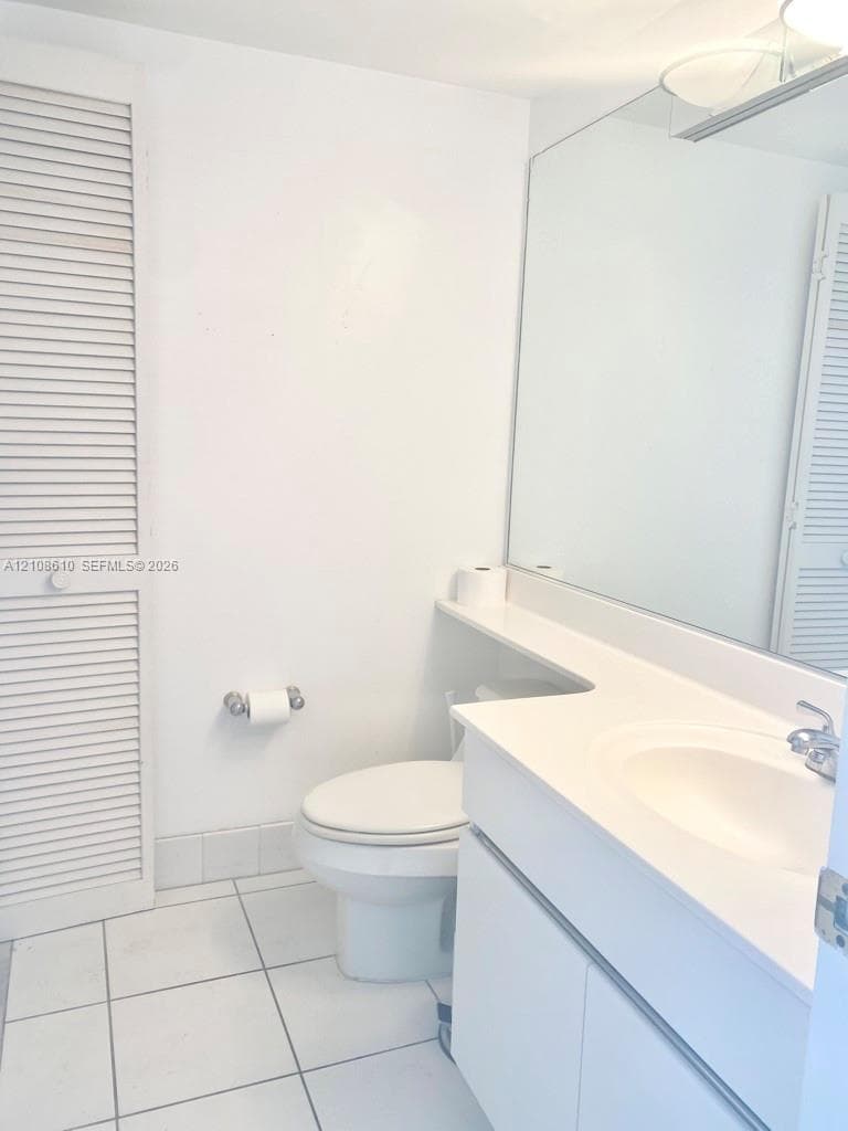 Bathroom featuring a white vanity cabinet, solid-surface countertop with an integrated sink, full-width vanity mirror, louvered closet doors, and ceramic tile flooring