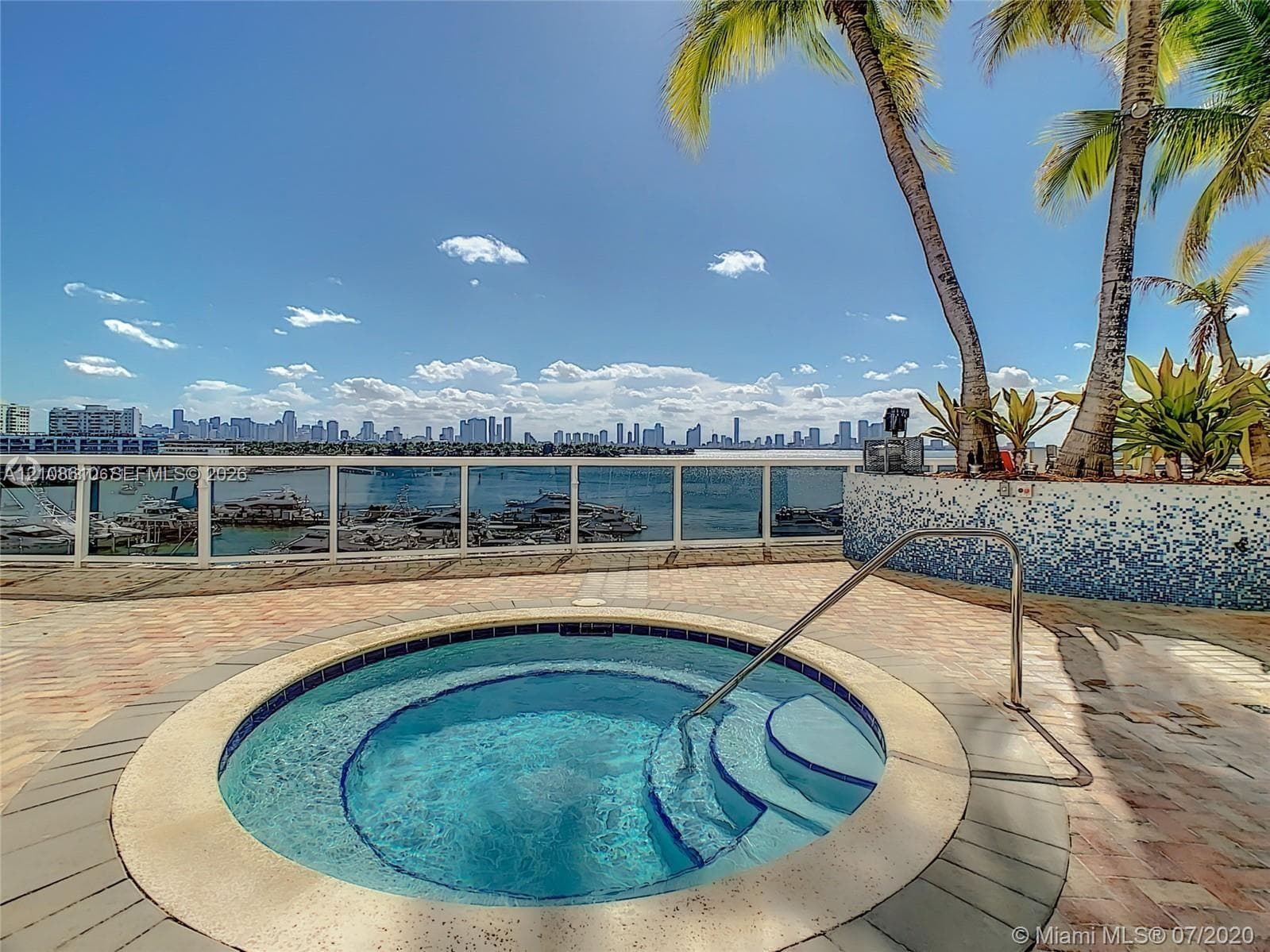 Outdoor spa featuring tiled surround, stainless steel handrail, paver decking, glass panel railing, and water-facing position