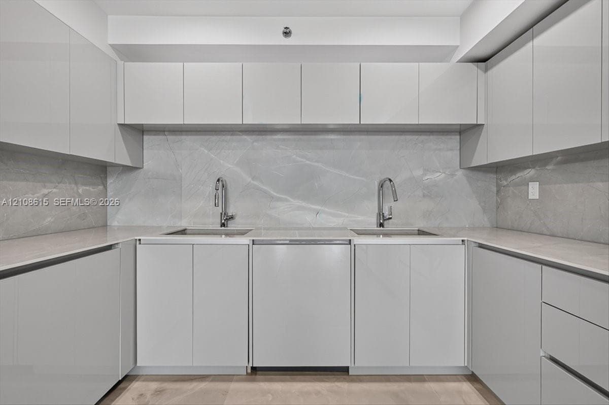 Glossy cabinetry, stone slab backsplash, dual undermount sinks, gooseneck faucets, and light-toned countertop surfaces