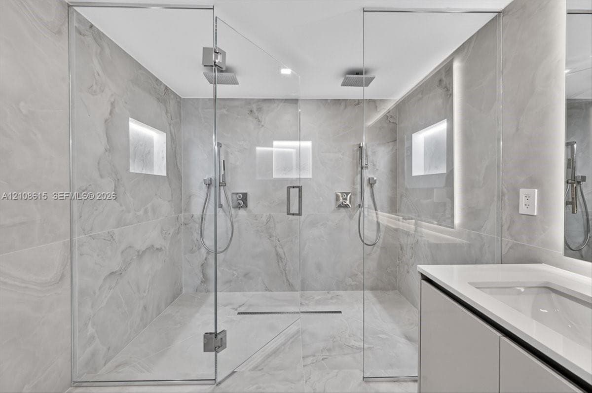 Walk-in shower featuring floor-to-ceiling stone-finish tile, dual ceiling-mounted rainfall showerheads, handheld sprayers, recessed wall niches, and frameless glass enclosure