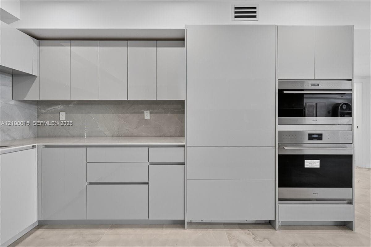 Modern kitchen featuring flat-panel cabinetry, stone-finish backsplash, neutral-toned countertops, and integrated stainless steel wall ovens