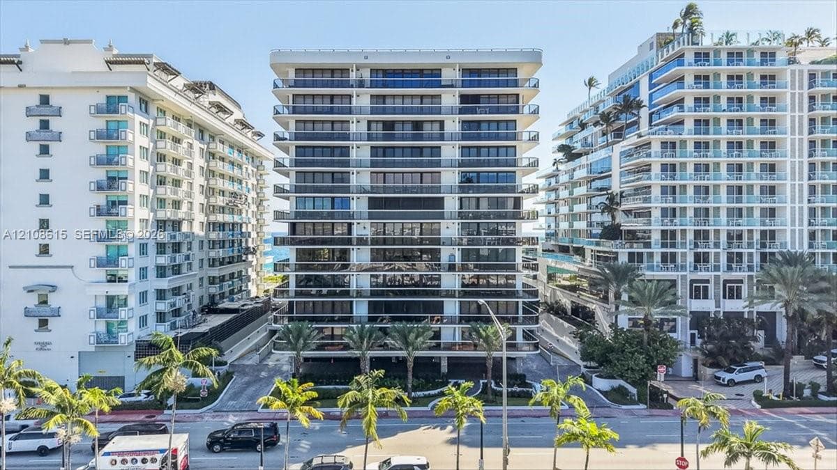 High-rise building exterior featuring tiered concrete balconies with glass railings, floor-to-ceiling windows, and mature palm trees
