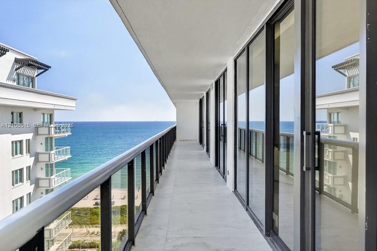 Expansive covered balcony featuring floor-to-ceiling glass doors, sleek metal railing, stone-finish flooring, and direct ocean exposure