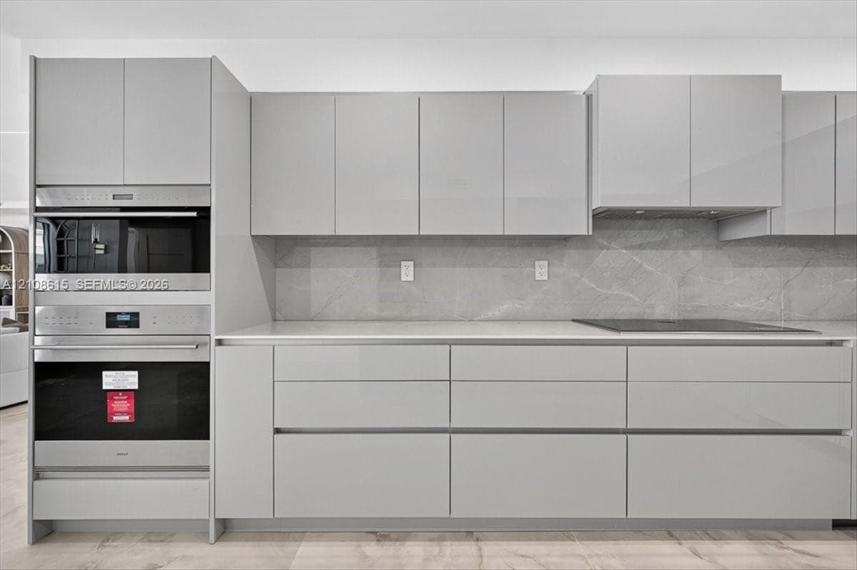 High-gloss cabinetry, stainless steel double wall ovens, expansive light-toned countertops, stone-finish slab backsplash, and integrated cooktop