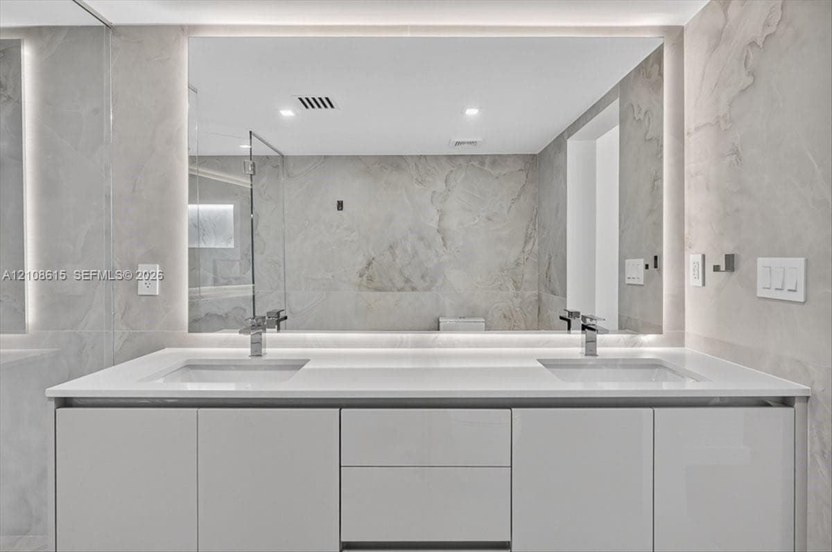 Modern dual-sink vanity featuring a solid-surface countertop, high-gloss cabinetry, integrated LED-backlit mirror, and polished metal fixtures against full-height stone-finish wall tile