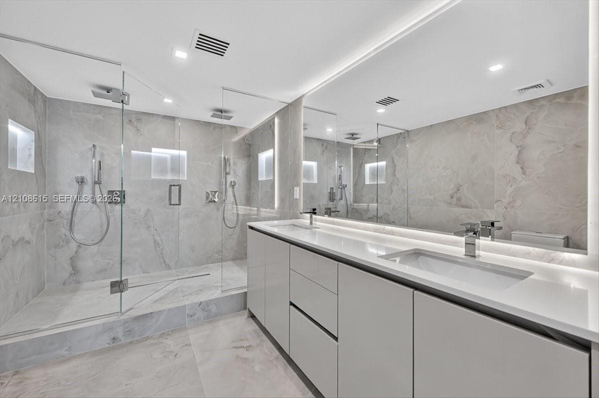 Bathroom featuring a walk-in shower with glass enclosure, large-format tile surround, dual vanity with undermount sinks, full-width mirror, and integrated lighting