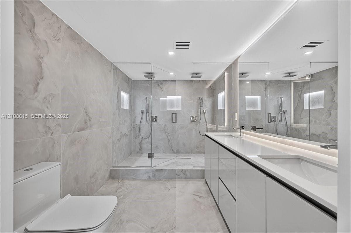 Dual-sink vanity with high-gloss cabinetry and solid surface countertop, walk-in glass-enclosed shower featuring dual rainfall showerheads and recessed wall niches, and floor-to-ceiling large-format tile surround