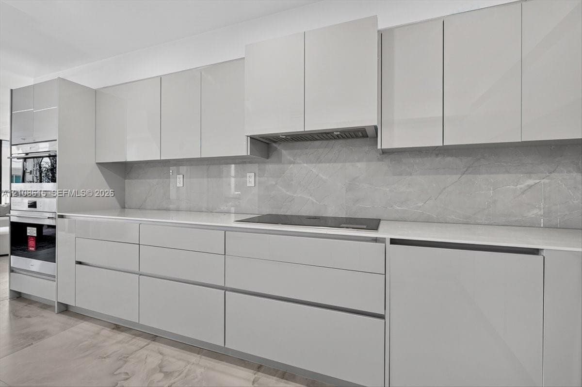 Sleek kitchen featuring high-gloss cabinetry, expansive stone-finish backsplash, built-in wall oven, flush-mount cooktop, and light-toned countertops