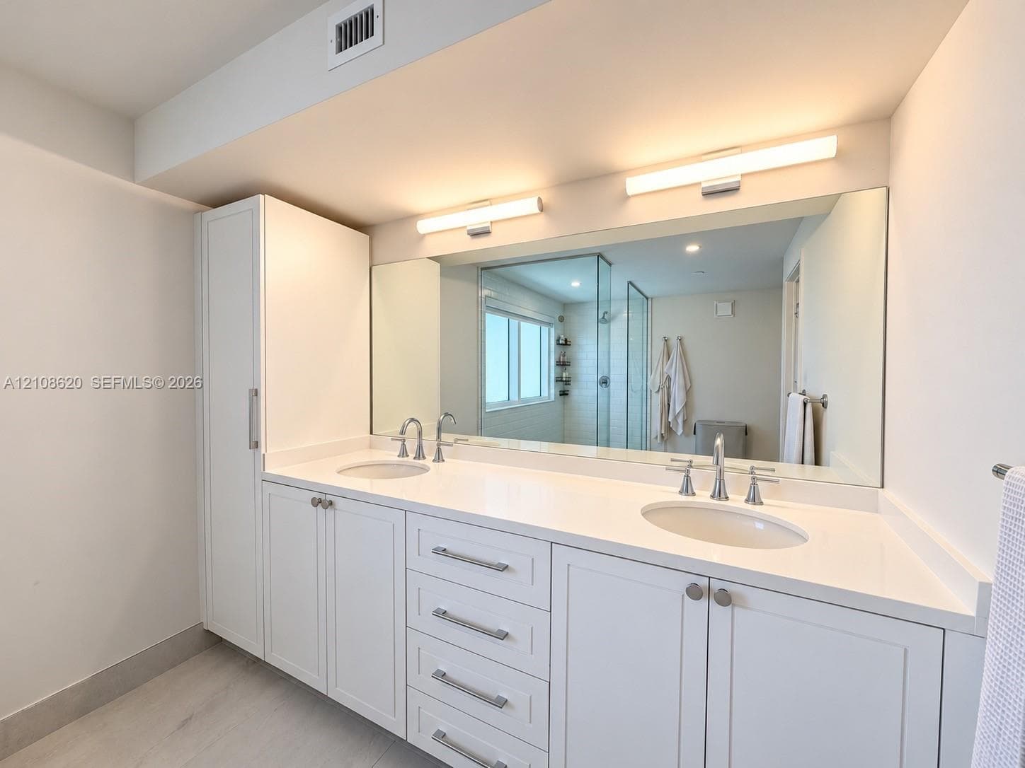 Dual-sink vanity featuring white shaker-style cabinetry, solid-surface countertop with integrated basins, brushed metal hardware, and full-width vanity mirror with dual wall-mounted light fixtures