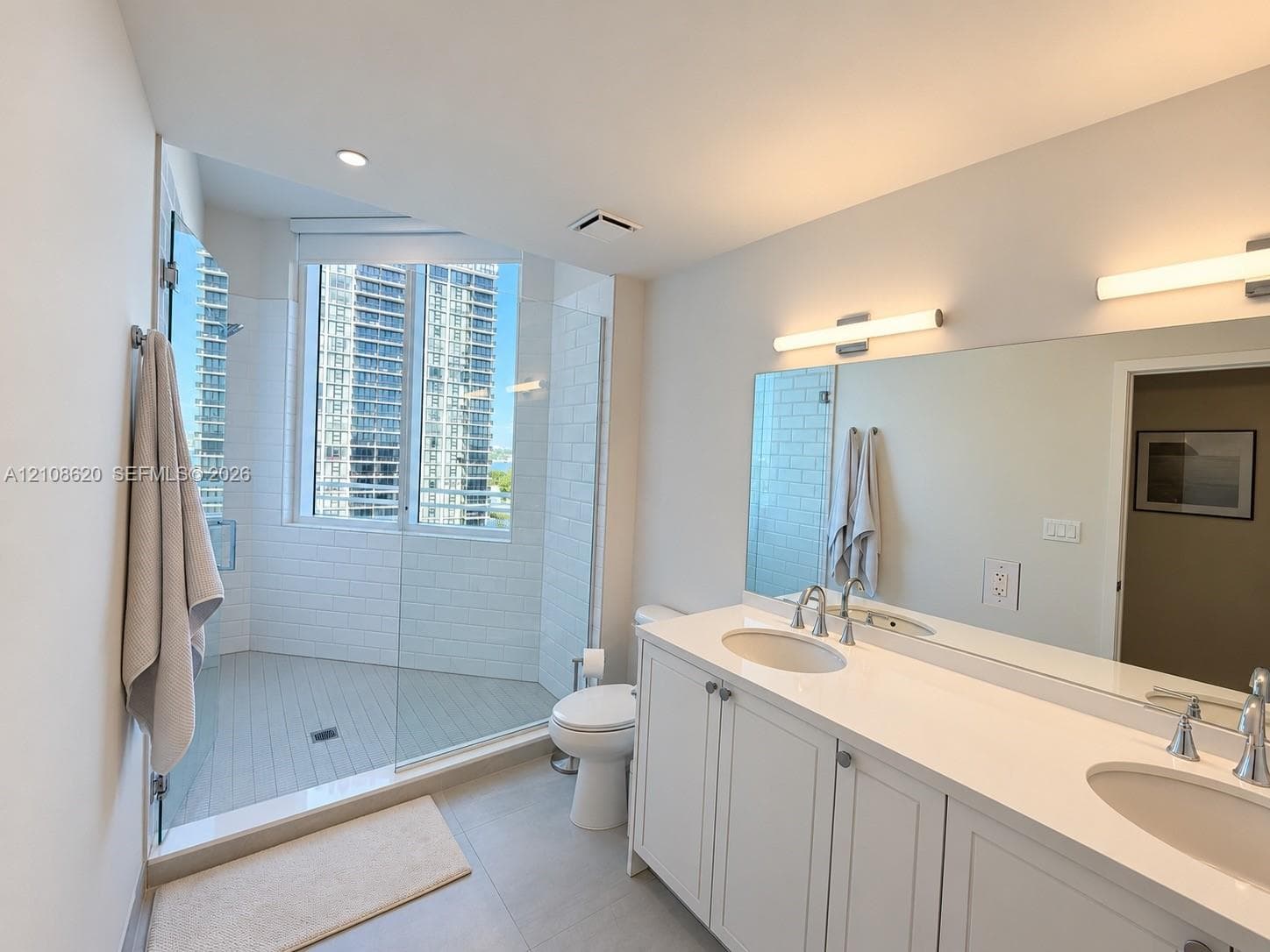 Bathroom featuring a dual-sink vanity with a solid-surface countertop, white shaker-style cabinetry, a walk-in shower with a glass enclosure and subway tile surround, and large-format floor tiling