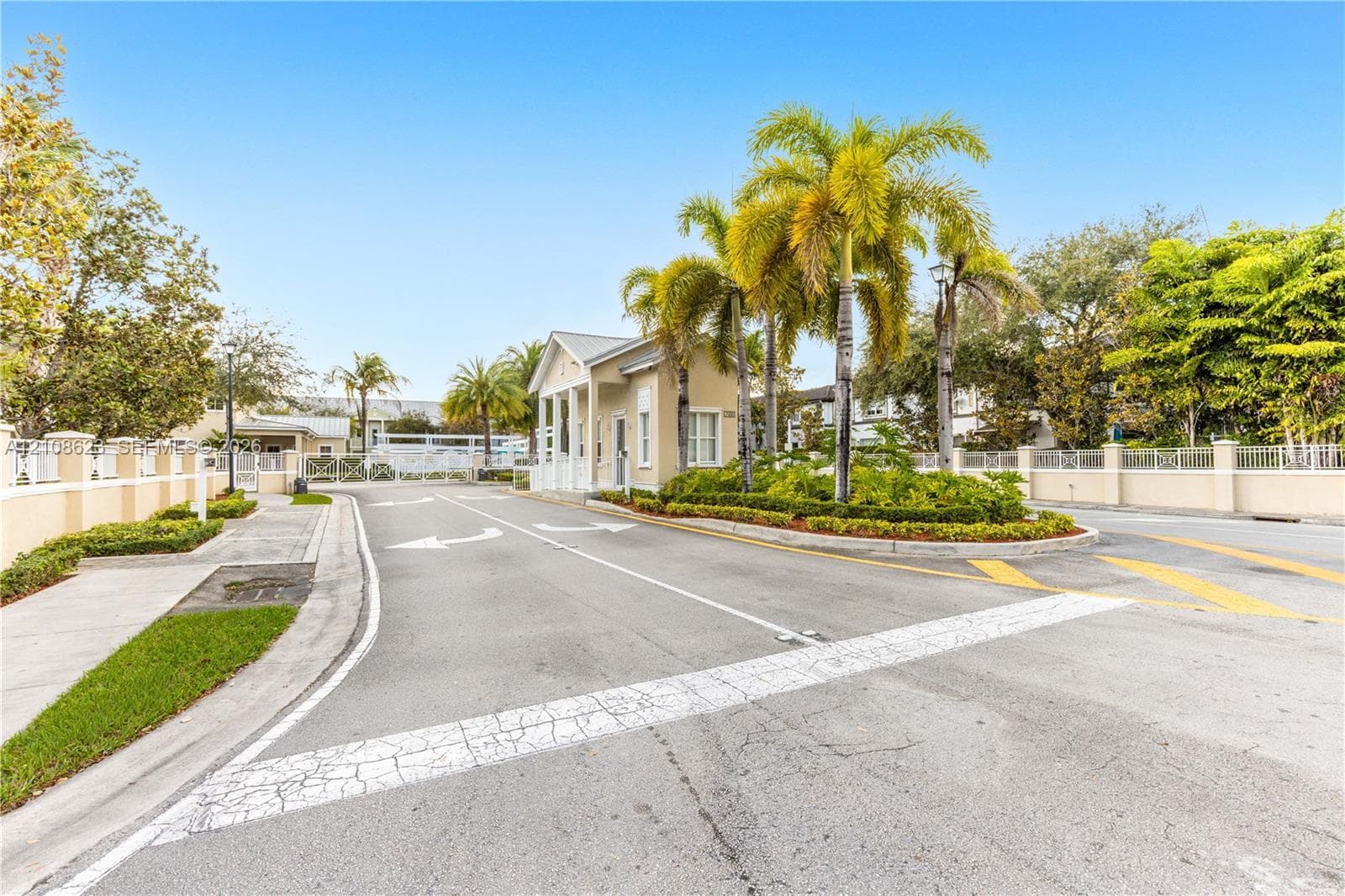 Gated property entrance featuring a security kiosk, concrete perimeter walls, paved roadways with directional markings, and mature palm landscaping