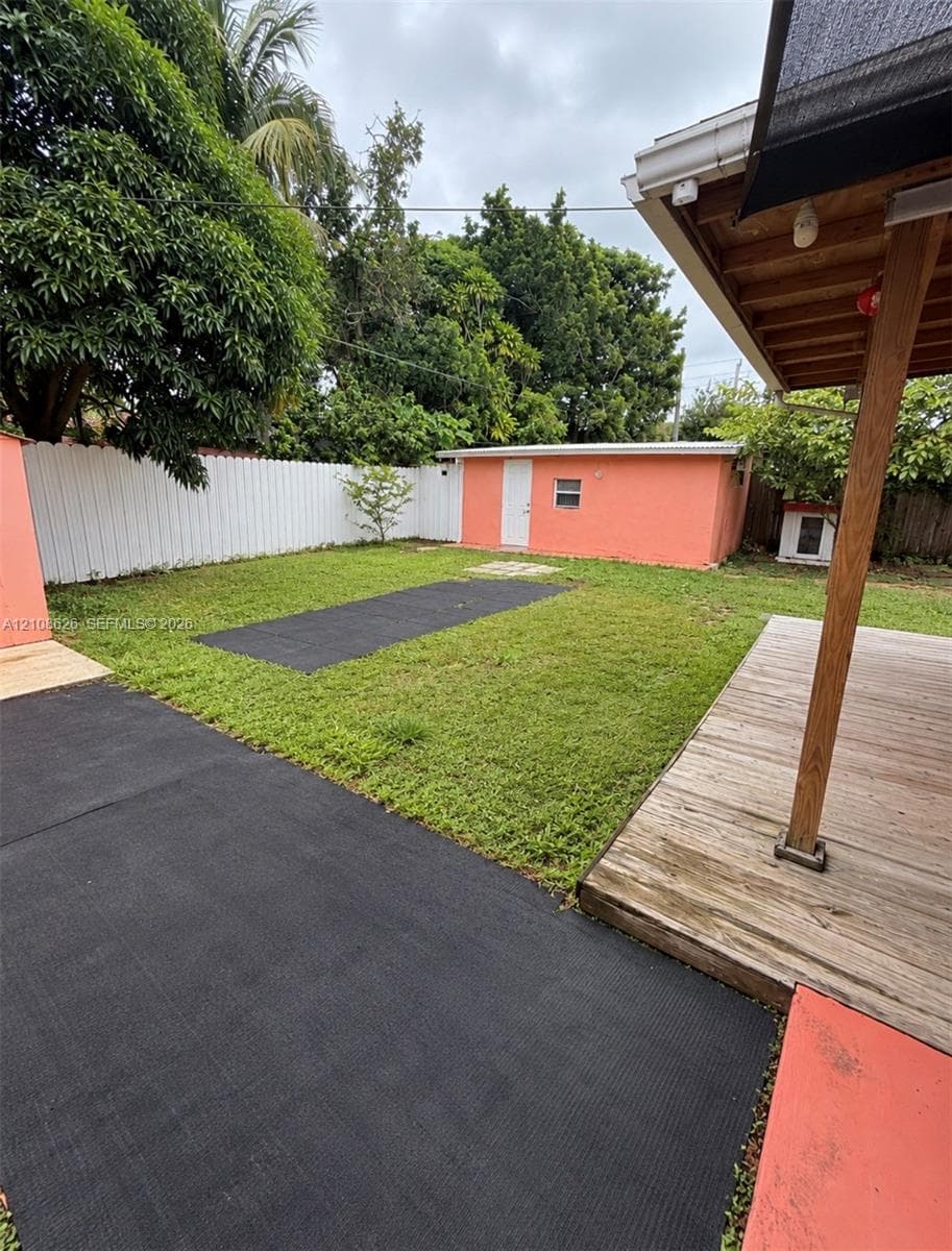 Backyard featuring a wood-plank deck with a covered area, a detached structure, a white privacy fence, and paved ground surfaces