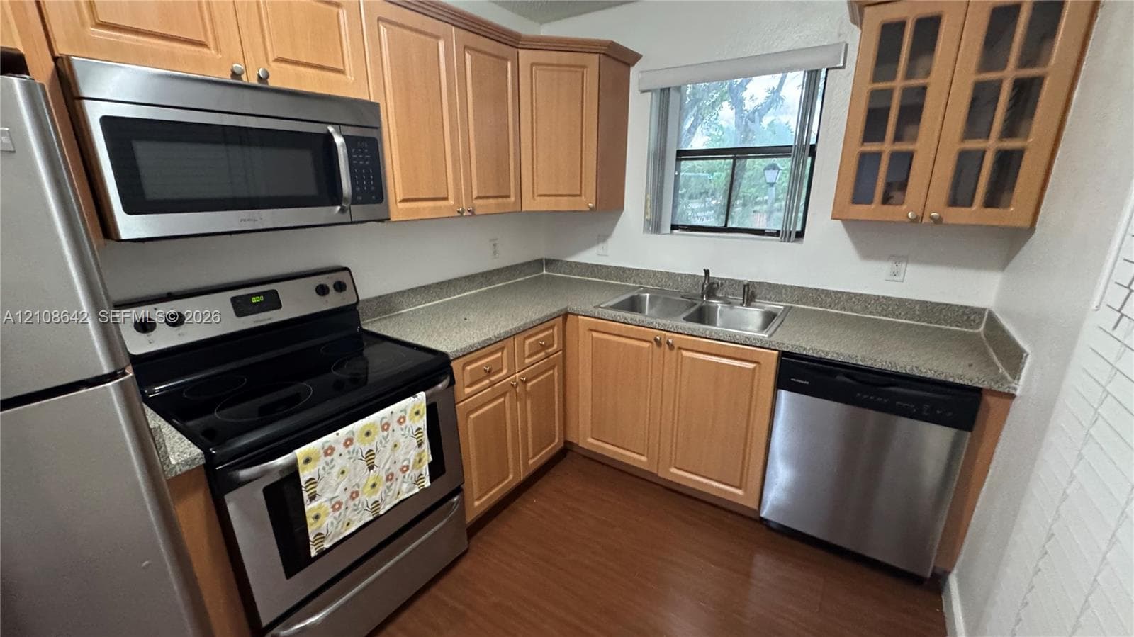 Kitchen featuring wood cabinetry, stainless steel appliances, speckled stone-composite countertops, a double-basin stainless steel sink, and wood-finish flooring
