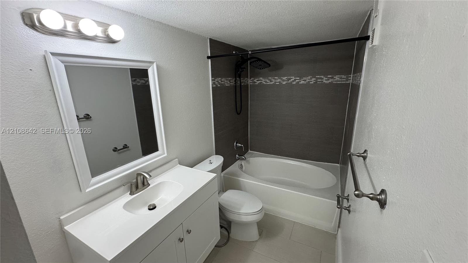 Bathroom featuring a white vanity cabinet with an integrated basin, a wall-mounted light fixture, and a soaking tub surrounded by dark tile with a decorative accent strip