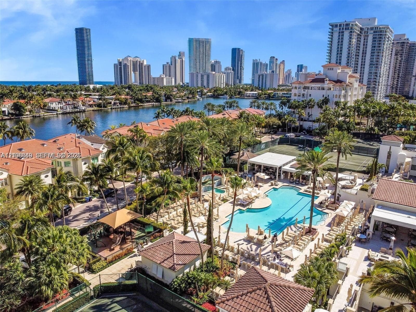 Waterfront community featuring an expansive swimming pool, professional-grade tennis courts, mature tropical landscaping, and tile-roof architecture