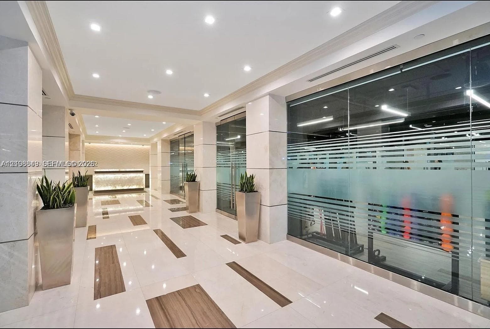 Polished stone flooring with inlaid wood-finish accents, large-format stone pillar cladding, glass-partitioned wall panels, recessed lighting, and a backlit stone-front desk