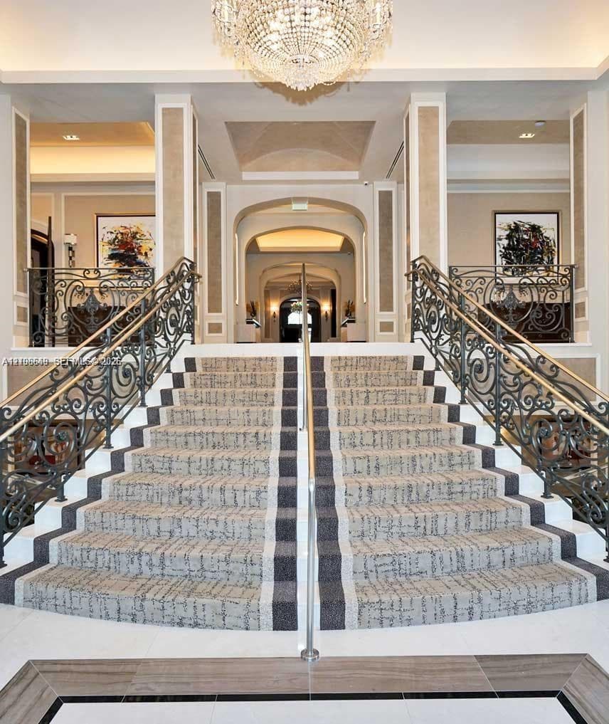 Grand dual staircase featuring intricate wrought iron railings, stone flooring with decorative inlay, and a central crystal chandelier under vaulted ceiling detailing