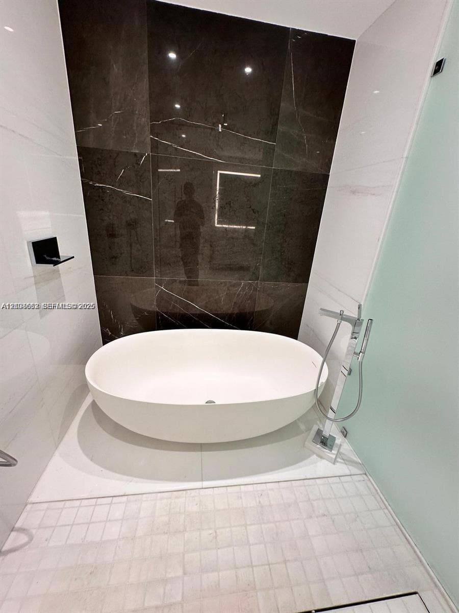 Freestanding oval soaking tub featuring a chrome floor-mounted faucet with a handheld sprayer, set against a dark large-format tile accent wall and white wall tile with a glass partition