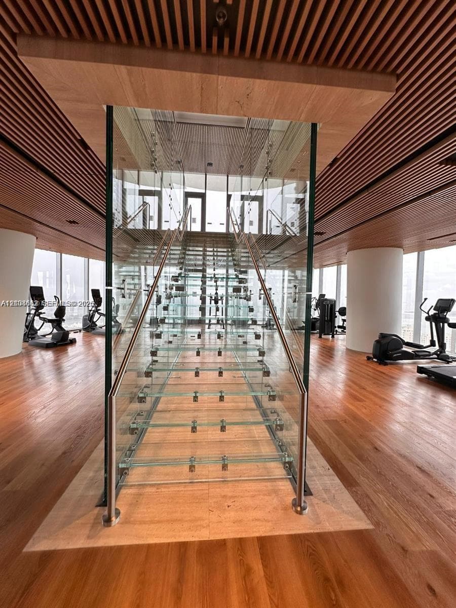 Staircase featuring glass treads and stainless steel railings, encased in a glass structural surround, complemented by a slatted wood ceiling, wood-finish flooring, and floor-to-ceiling windows