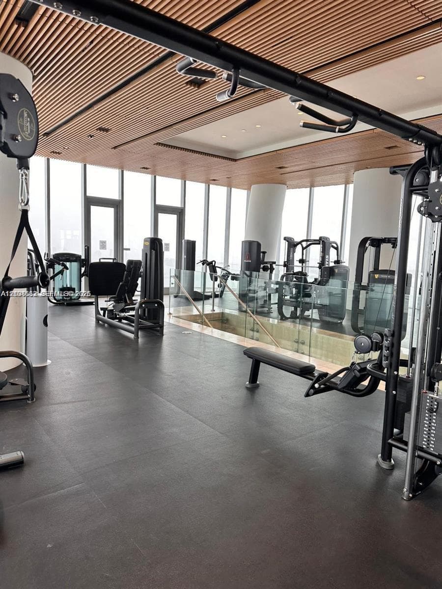 Fitness center featuring floor-to-ceiling windows, linear wood-slat ceiling, dark resilient flooring, and glass partition railings