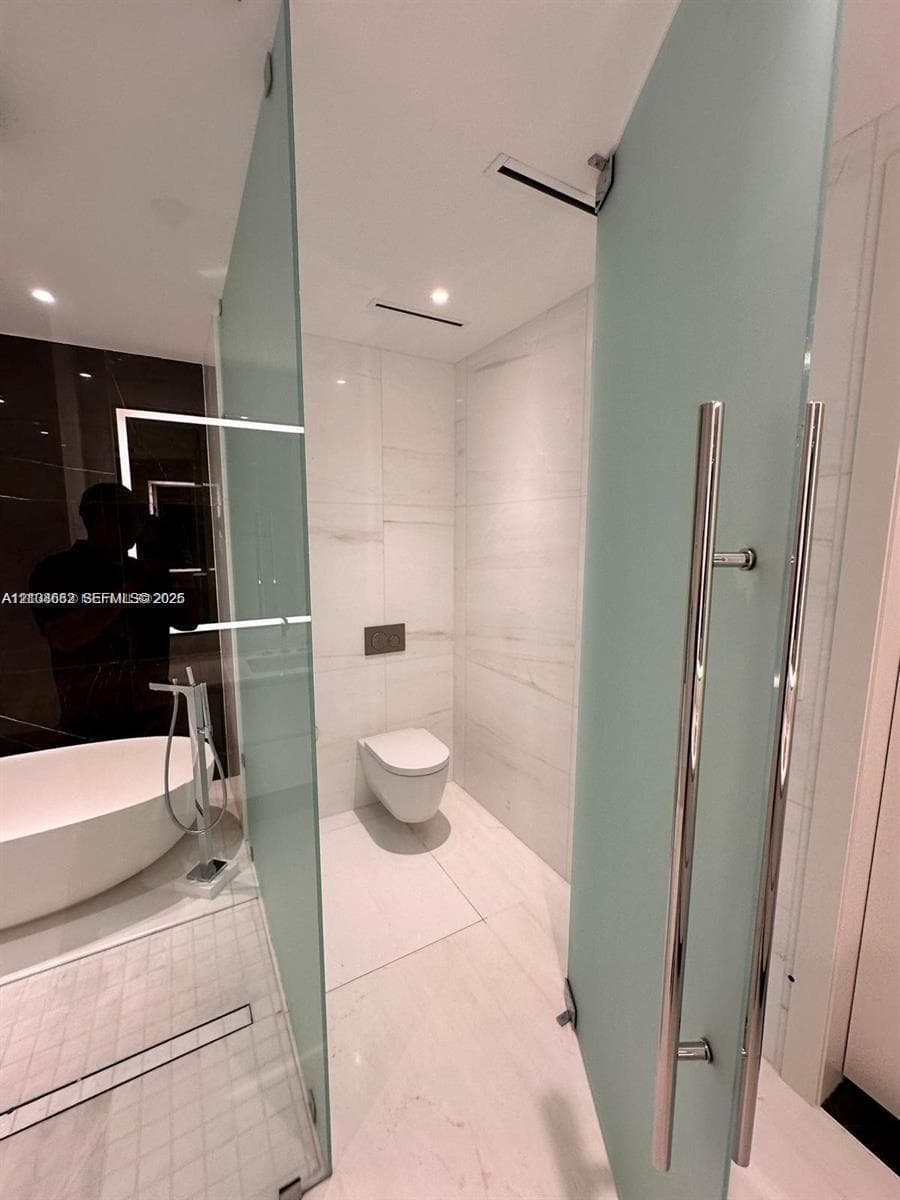 Freestanding soaking tub with floor-mounted chrome faucet, wall-hung toilet, large-format tile flooring, and frosted glass partition with polished chrome hardware