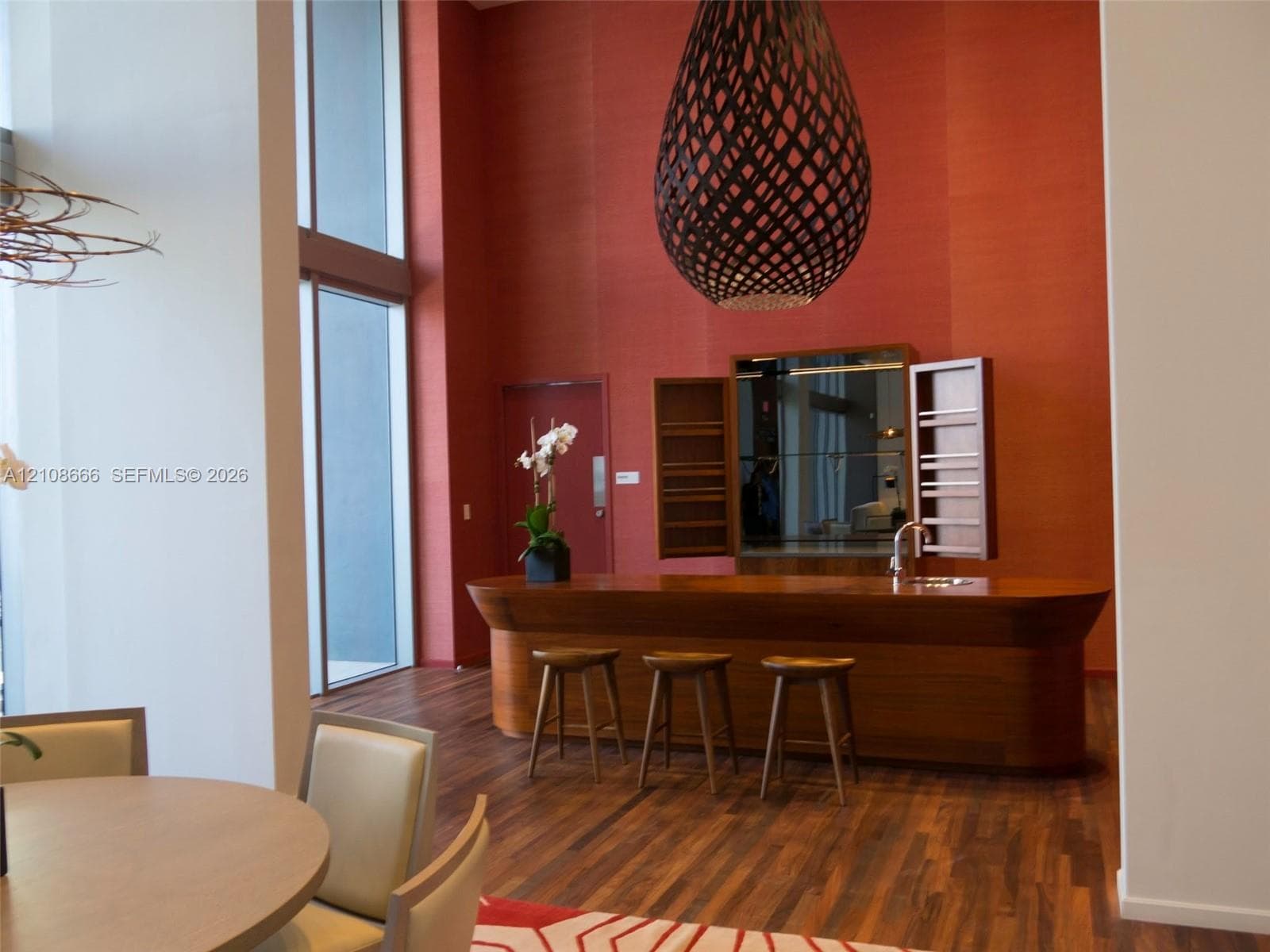 Double-height windows, wood-finish flooring, custom wood-paneled bar with integrated sink, and an oversized decorative pendant light fixture