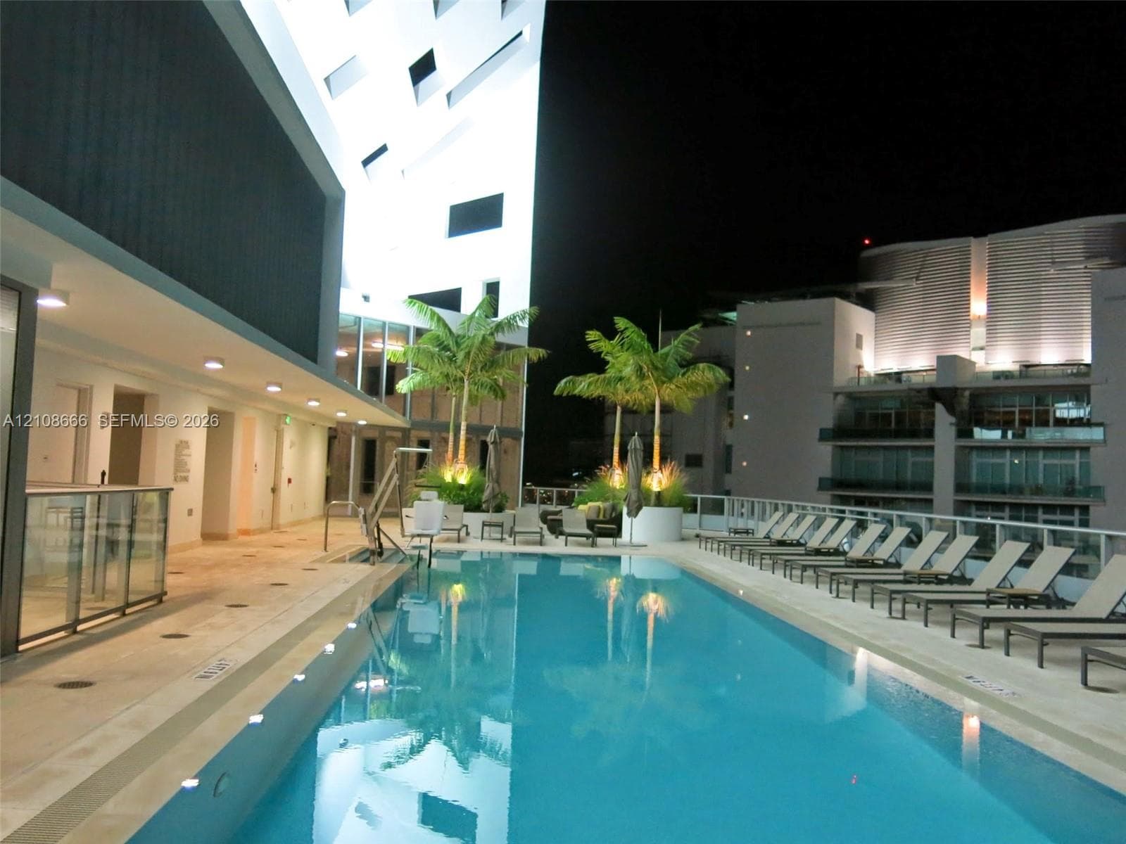 Rectangular swimming pool featuring a light-toned deck, integrated pool lift, recessed lighting, and palm tree planters