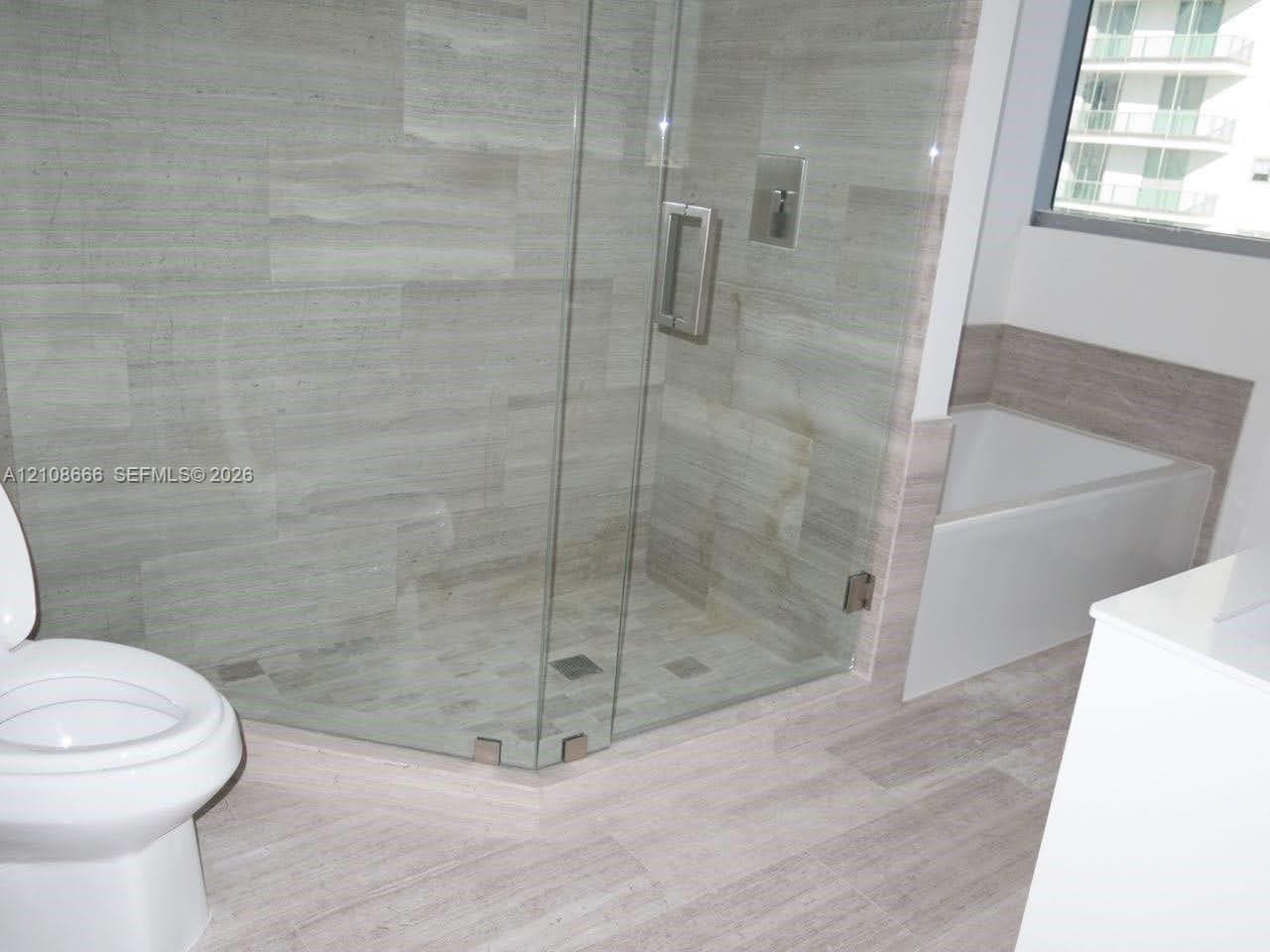 Walk-in glass-enclosed shower featuring large-format stone-finish tile surround and floor, deep soaking tub with tile surround, and neutral wood-finish flooring