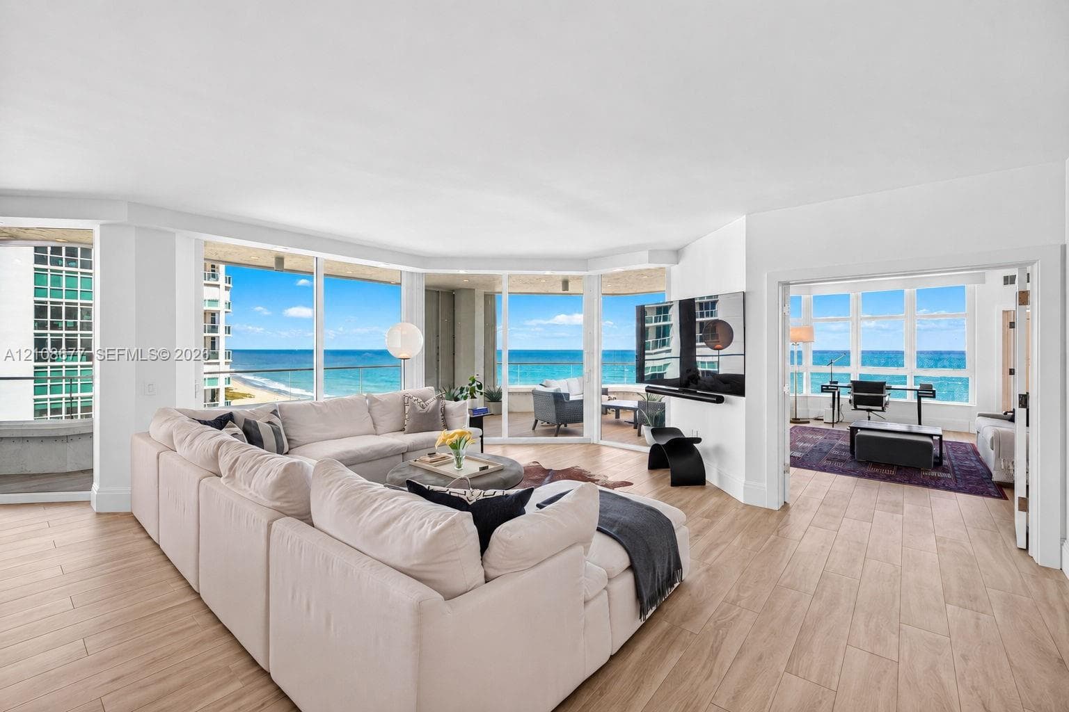 Expansive living area (and bedroom to the right, currently being used as a den) w/ hurricane impact rated floor-to-ceiling windows, wood look plank porcelain flooring, recessed lighting, and a wrap-around balcony with panoramic ocean views. Light filtering remote operated shades with Bandalux