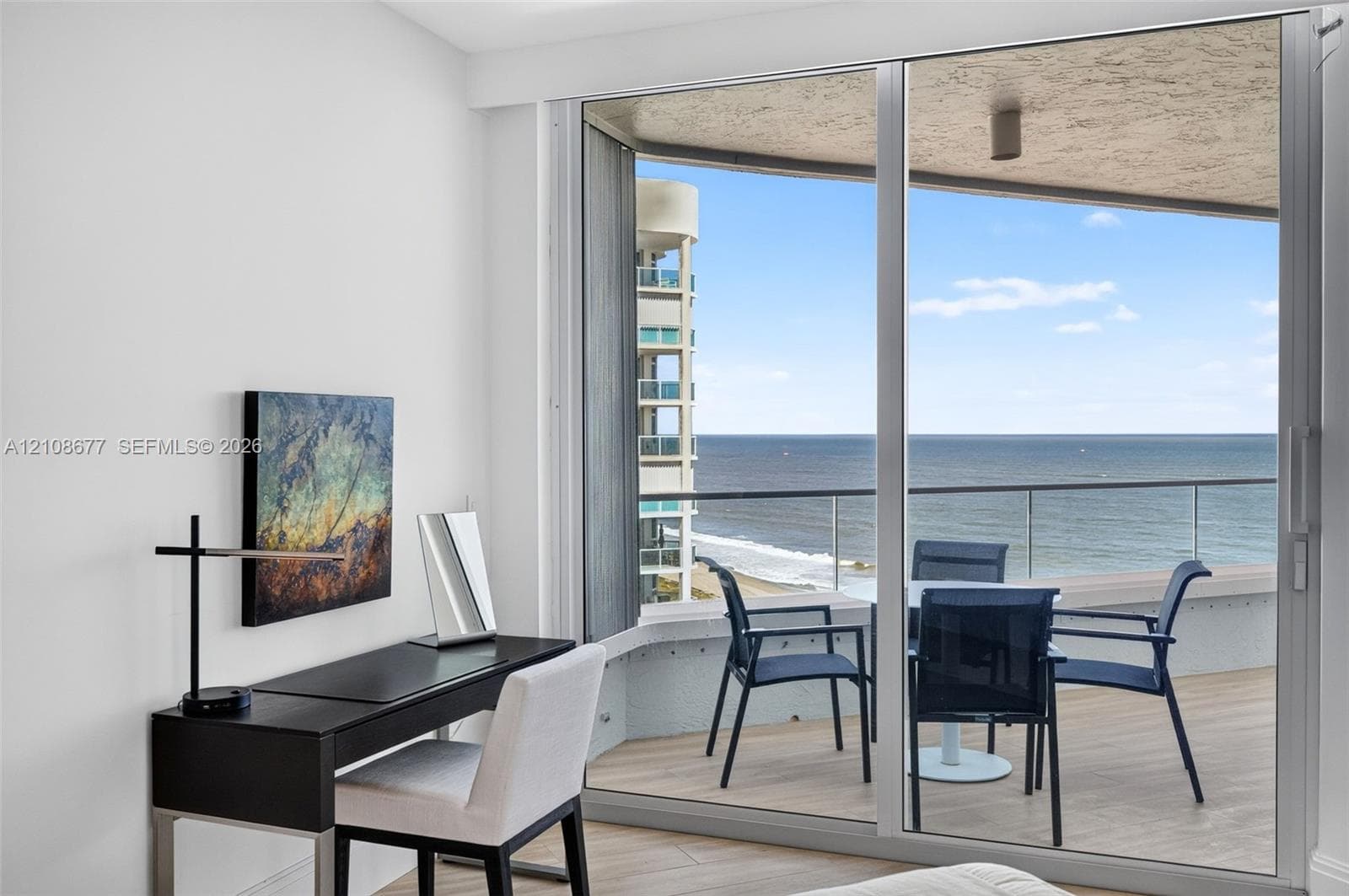 Full-height sliding glass doors opening to a private balcony with glass railing, wood look plank porcelain flooring, and ocean scenery. Double layered (light filtering and room darkening) remote operated window shades by Bandalux