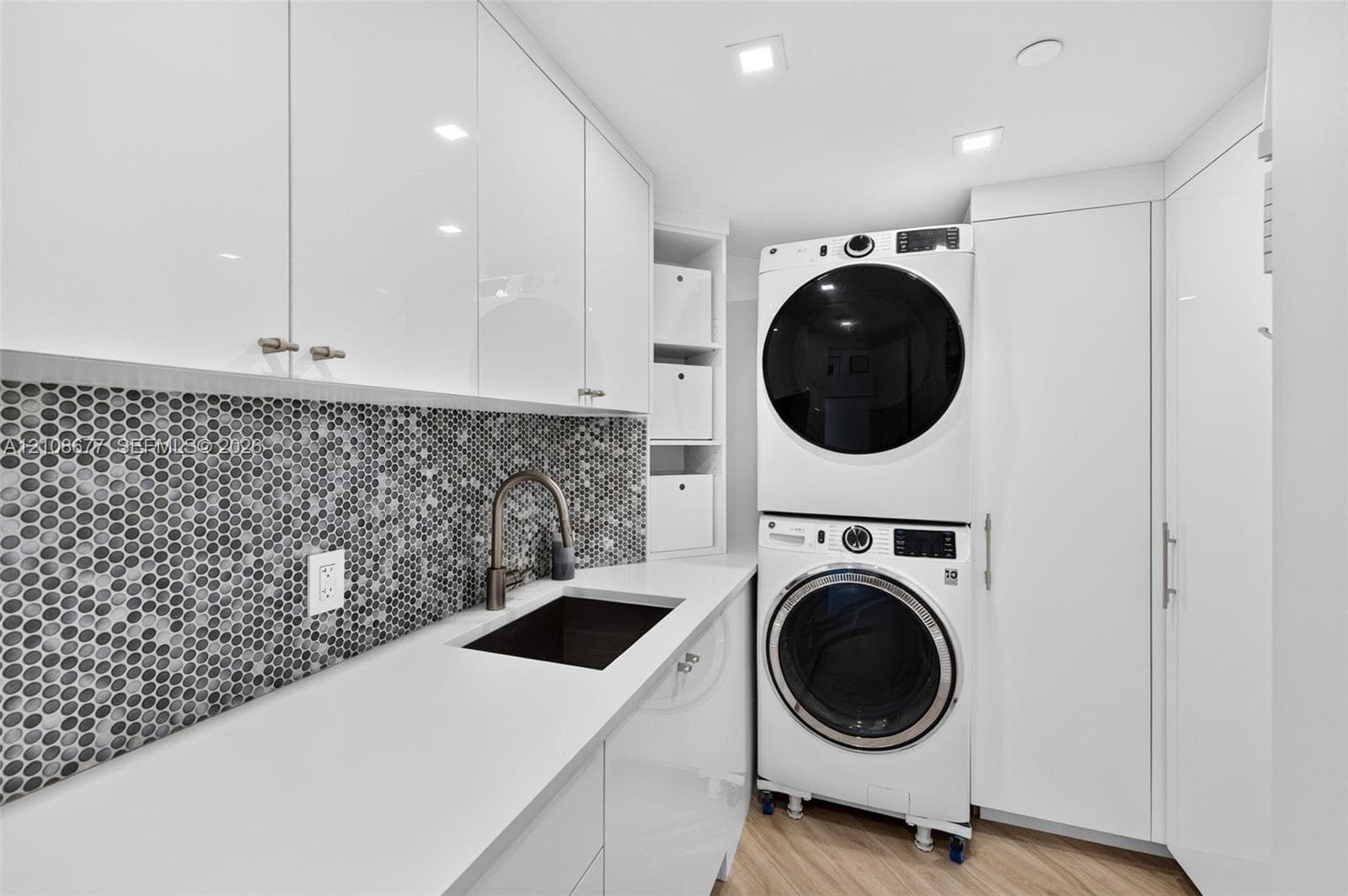 Expanded Laundry room featuring high-gloss cabinetry, quartz counters, mosaic penny-tile backsplash, an integrated basin sink with a gooseneck faucet, and recessed lighting. Full sized washer and dryer and tall broom / vacuum closet on the right