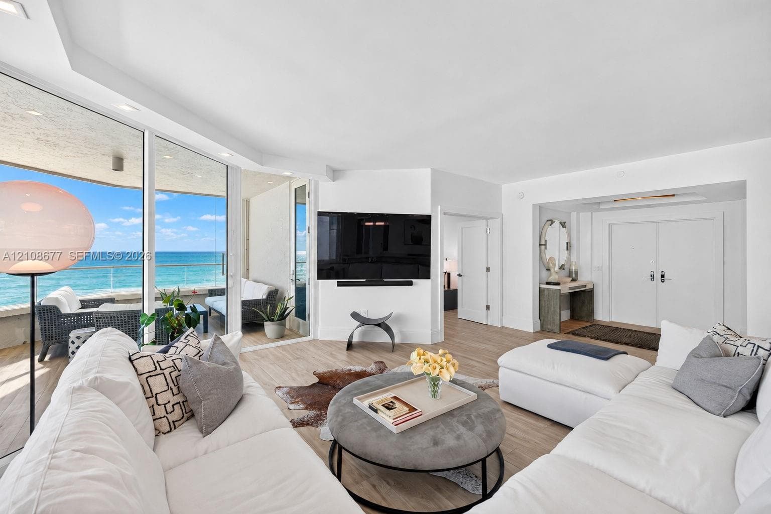 Living area featuring floor-to-ceiling glass panel walls, wood look plank porcelain flooring, integrated lighting, and a glass-railed balcony for more ocean views. TV wall was expanded to accommodate a larger TV. Light filtering remote operated shades with Bandalux