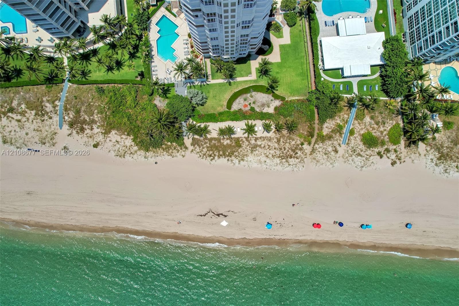 Aerial perspective of a multi-story residential building located on the coastline, featuring an on-site tennis court, swimming pool, and direct proximity to the ocean and sandy beach