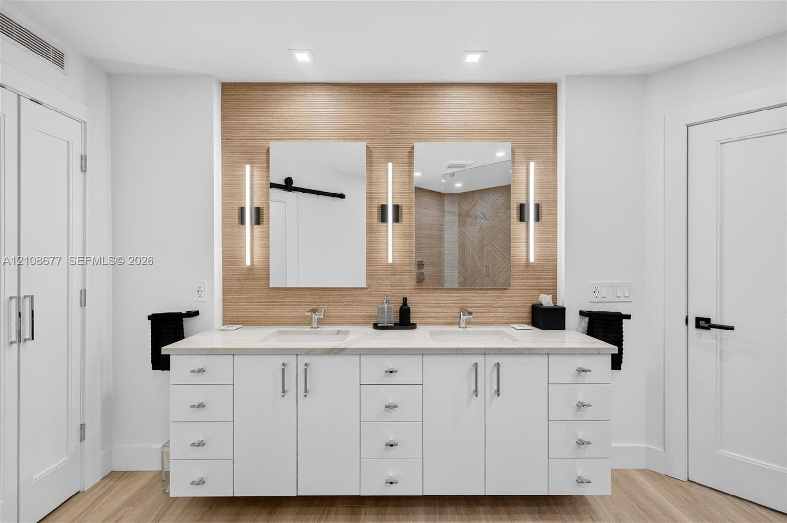 Dual-sink custom vanity featuring white cabinetry, light-toned stone counter, warm wood inspired wall tile and wall-mounted vertical sconces