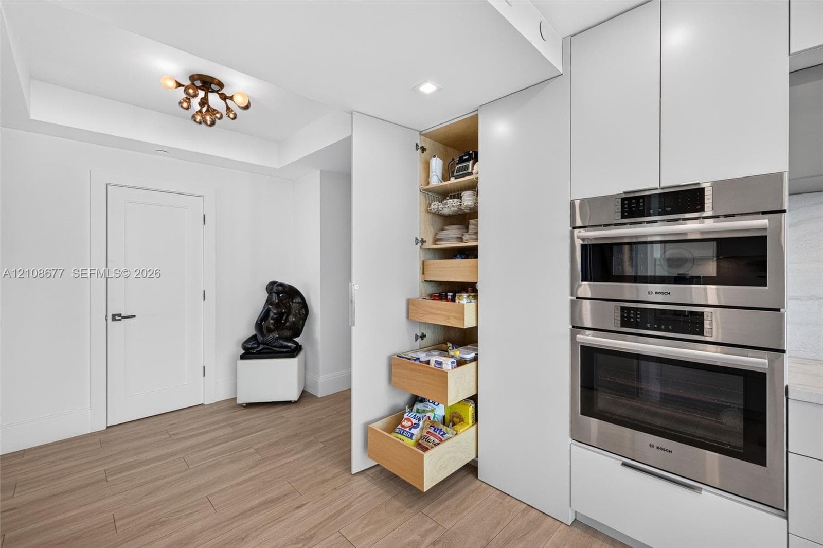 Custom cabinets featuring a floor-to-ceiling pull-out pantry with wood pull out and soft-close tongue & groove drawers, integrated stainless steel Bosch ovens, and wood look plank porcelain flooring. Bosch double oven w/ convection, microwave & speed oven