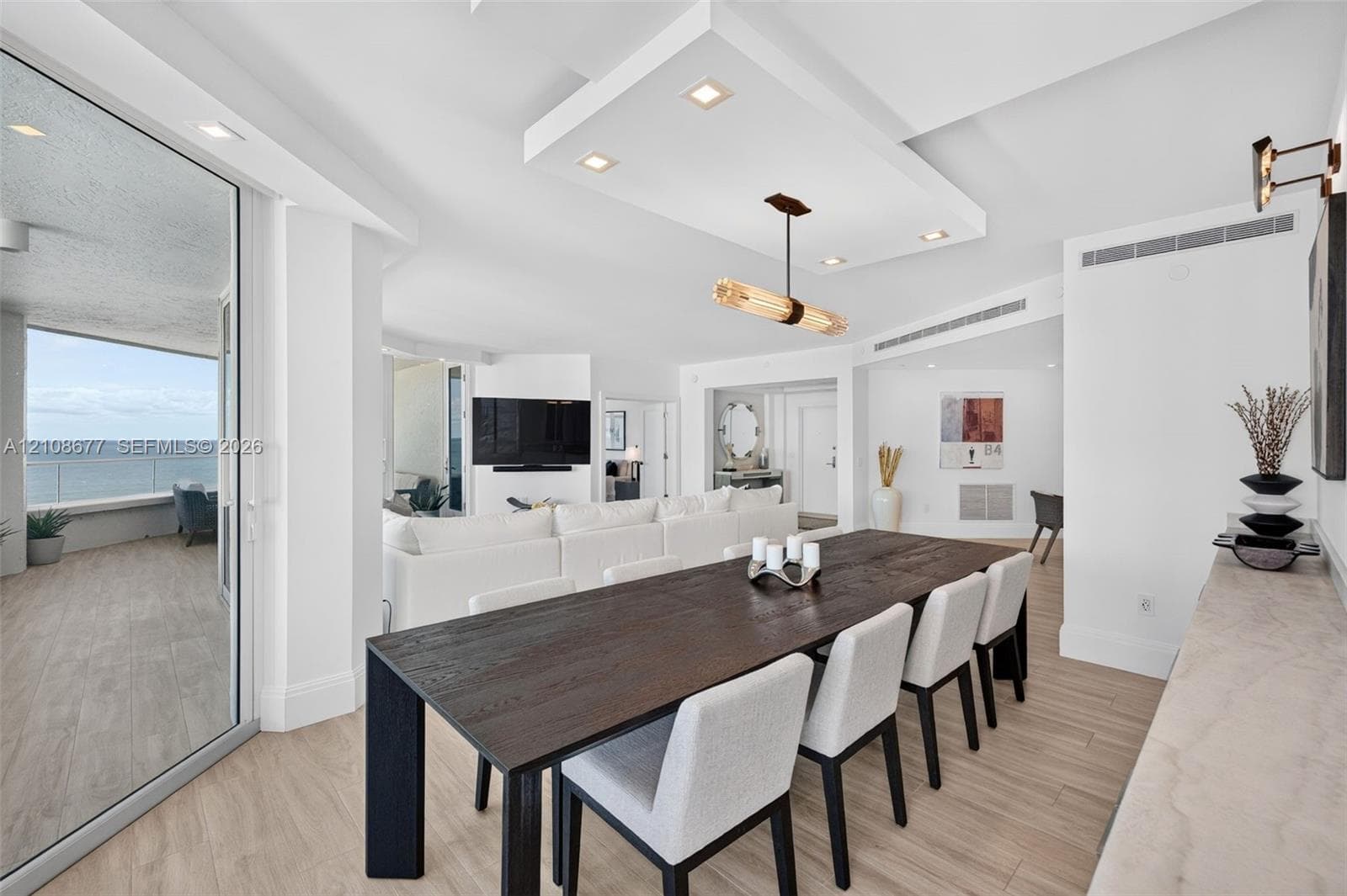 Open-concept interior featuring wood look plank porcelain flooring, recessed lighting, a custom architectural ceiling, a modern linear chandelier, and floor-to-ceiling glass doors opening to an exterior balcony from dining area w/ built in custom buffet for extra storage. Sliders lead again to the balcony for entertaining