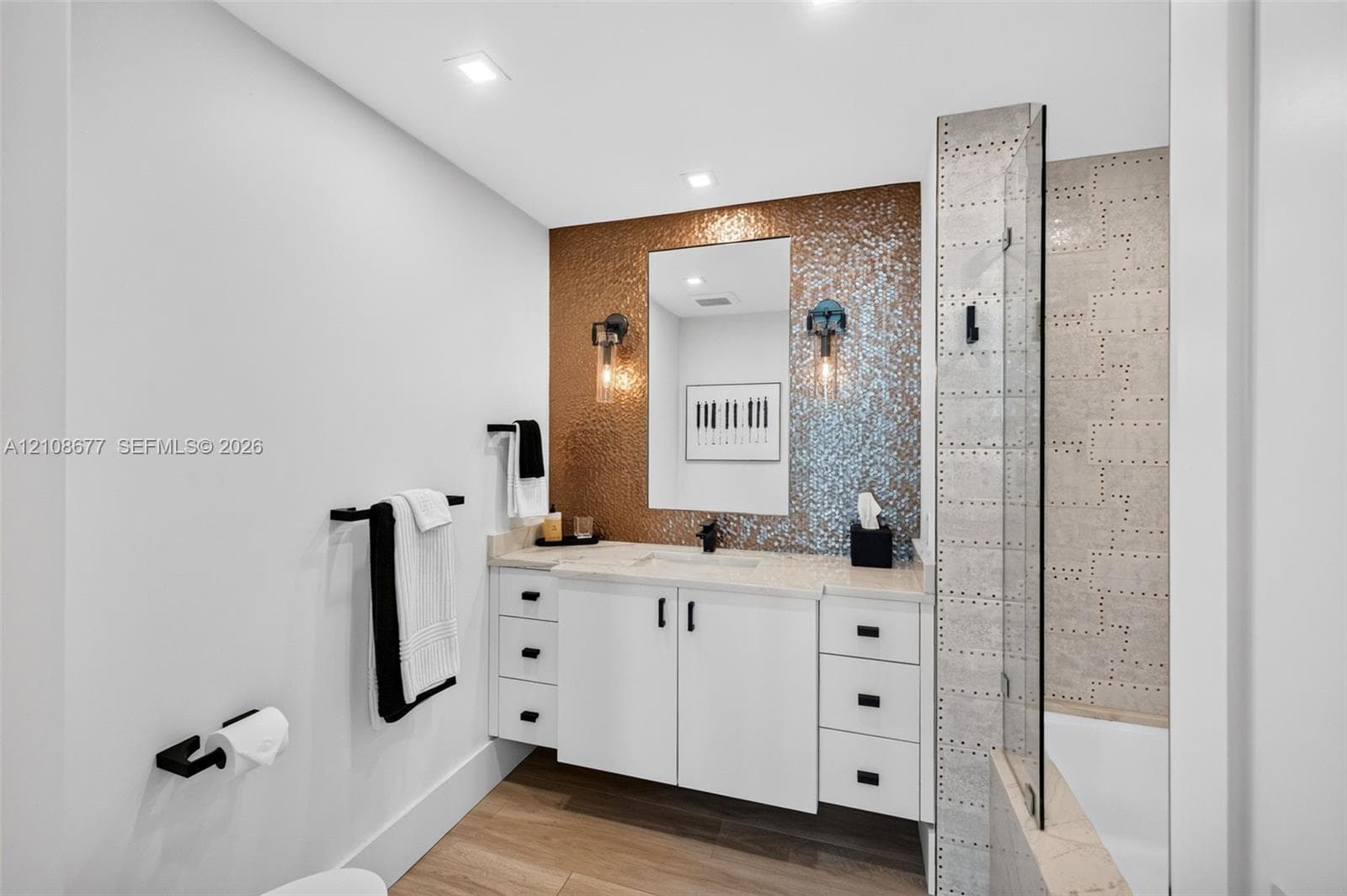 Bathroom w/ custom wall hung white vanity with matte black hardware, metallic mosaic tile accent wall, recessed lighting, and a glass-enclosed shower with custom industrial-inspired wall tile