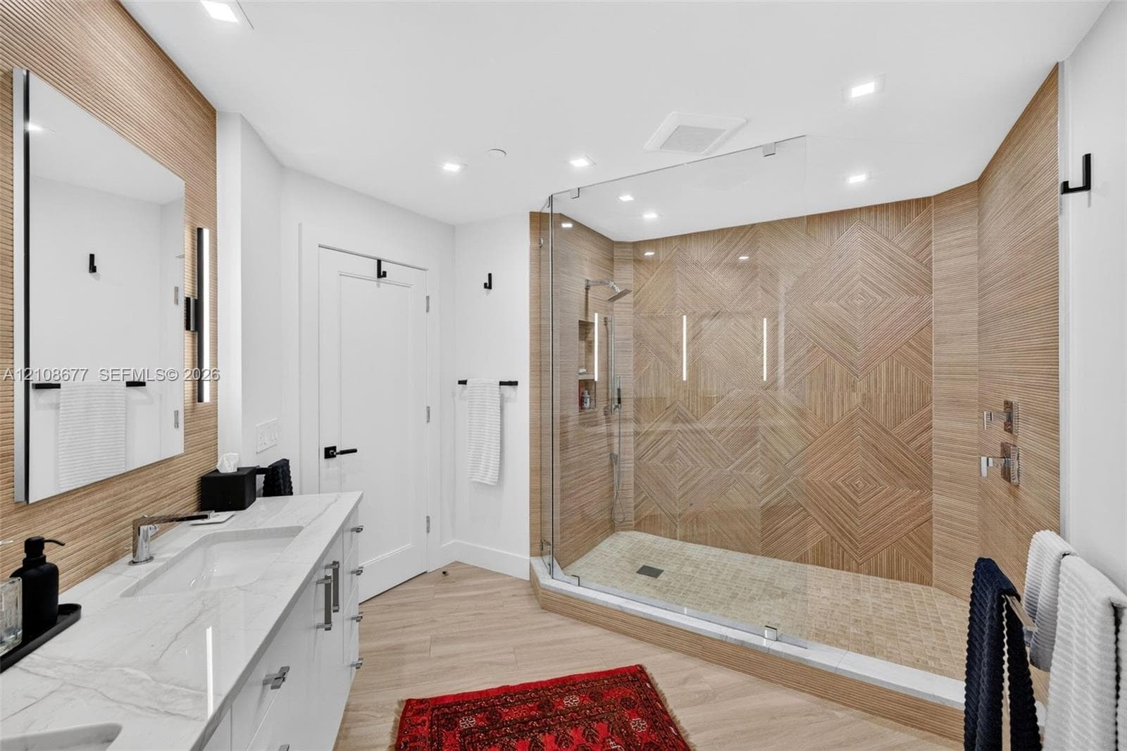 Modern bathroom featuring a dual-sink vanity with stone counters, a glass-enclosed shower with geometric tile work and built-in shelving, and wood-look wall porcelain