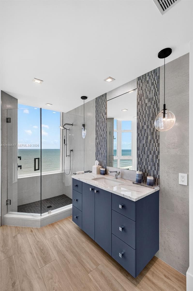 Custom navy-blue vanity with a stone countertop, glass-enclosed shower with tiled flooring, wood look plank porcelain flooring, and accent tile backsplash